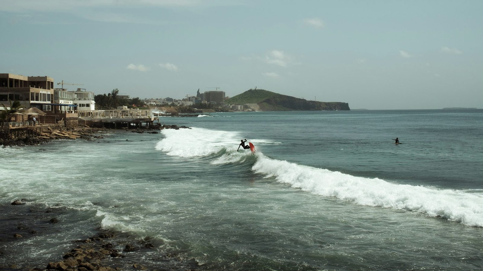 A surfer rides a breaking wave beside a rocky shore with houses and a headland beyond