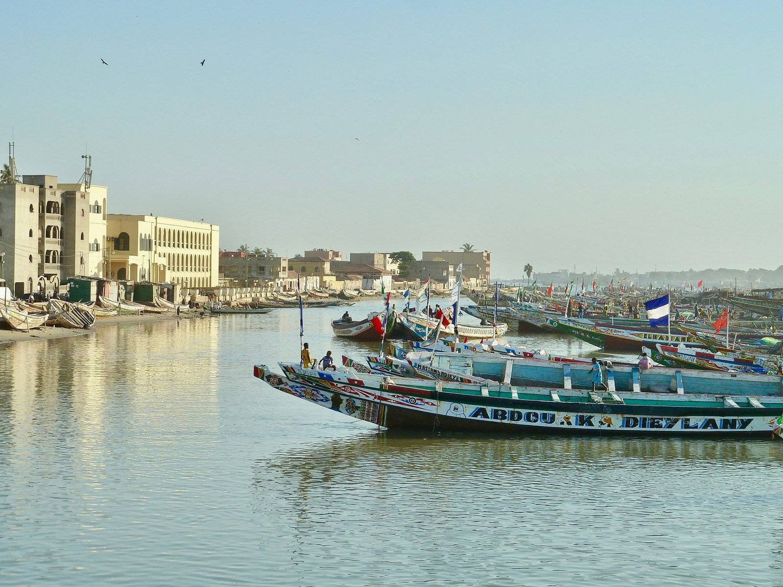 Long painted wooden fishing boats line a calm river below pale multi-story buildings