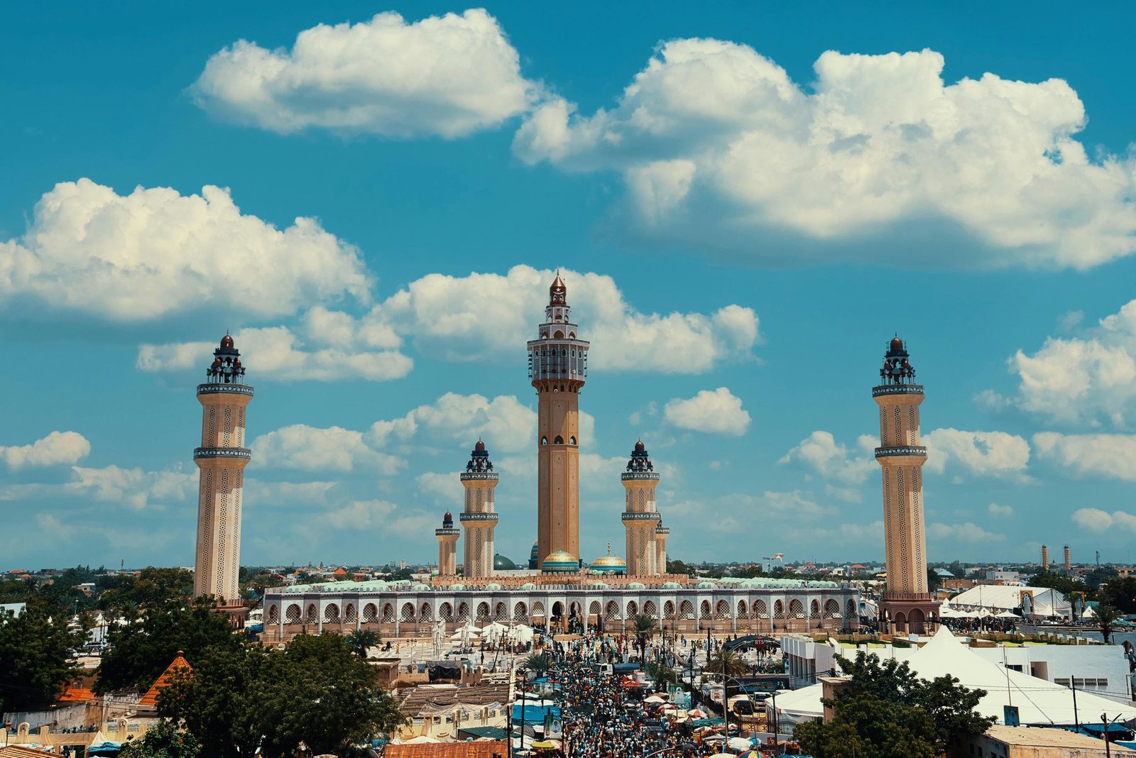 A mosque with a tall central minaret and four corner minarets rises above a crowded town under blue sky with white clouds