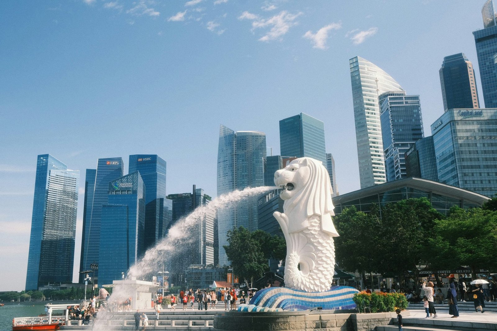 A white lion-headed fish statue spouts water into the bay in front of glass skyscrapers and a crowd of visitors