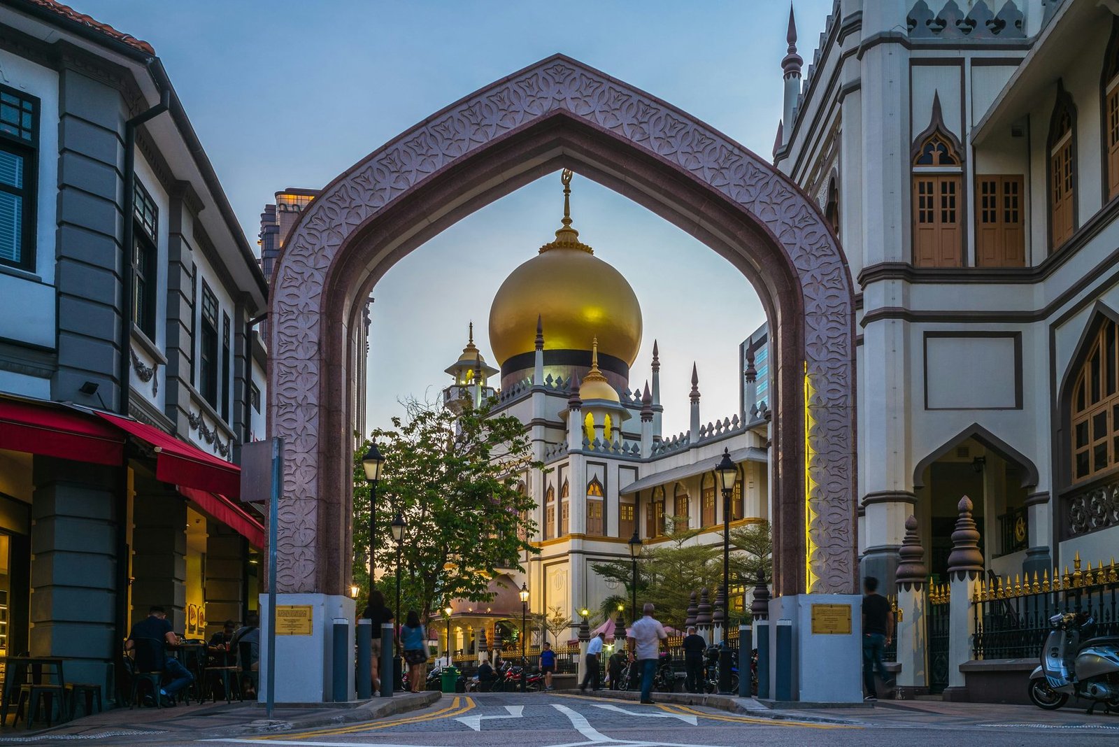 A pointed patterned arch frames a mosque with a large gold dome at the end of a street of shophouses