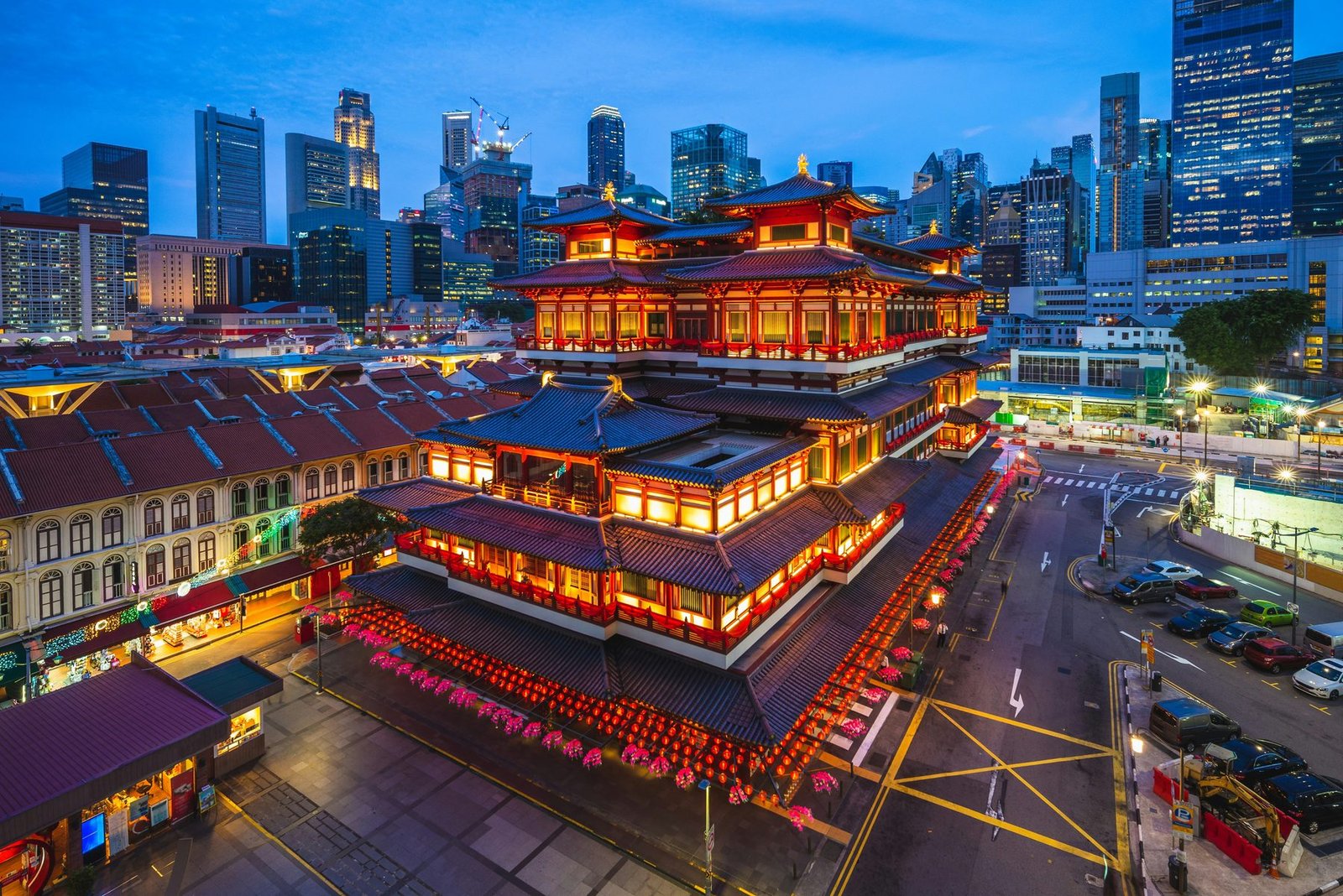 A red multi-tiered temple outlined in lights stands among low shophouses with office towers behind under a blue evening sky