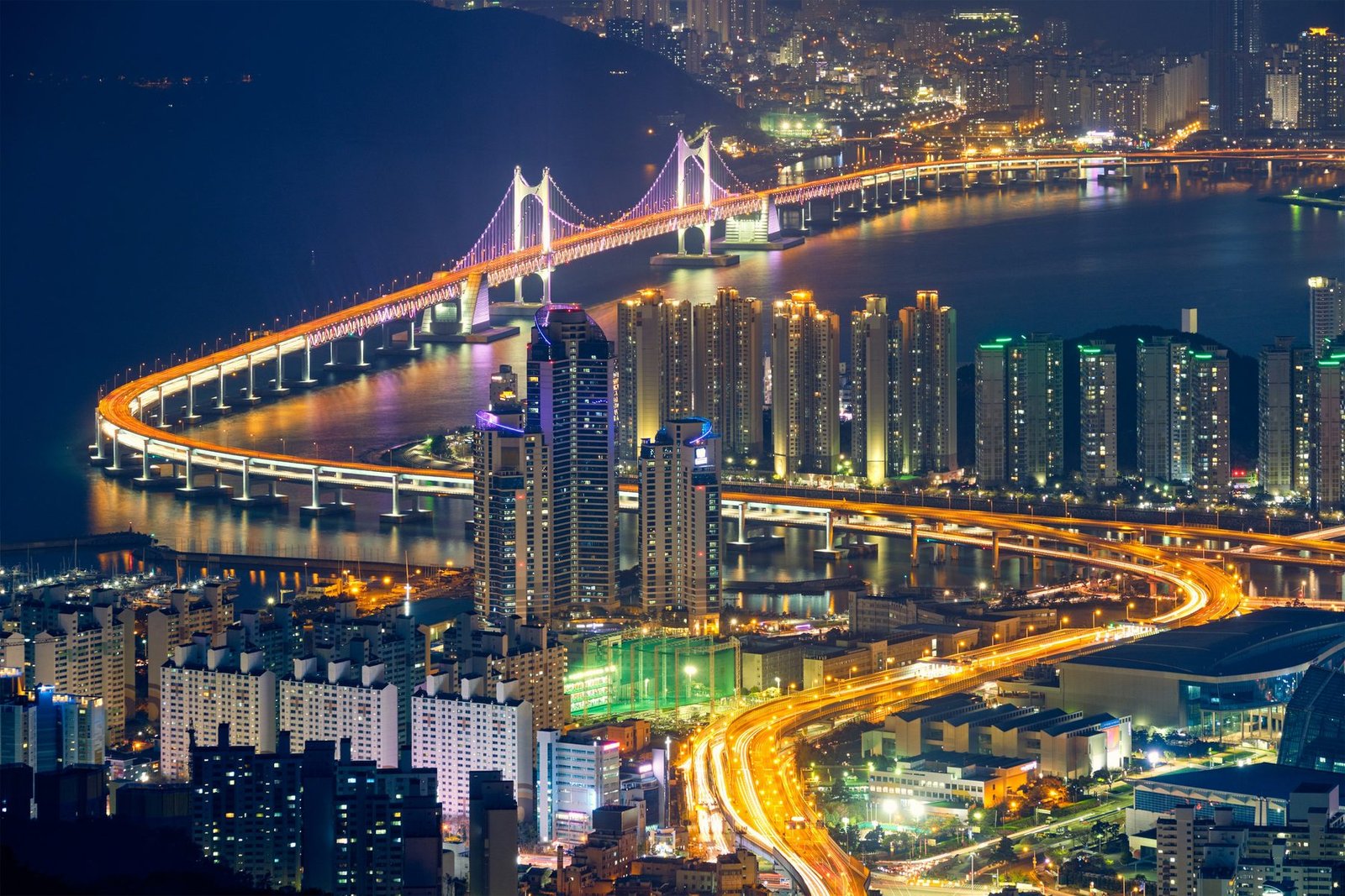 A lit suspension bridge curves across a dark bay past apartment towers, with orange light trails on the roads below, seen from above