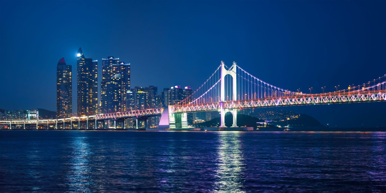 A suspension bridge lit in white and pink spans dark blue water with a cluster of towers at left