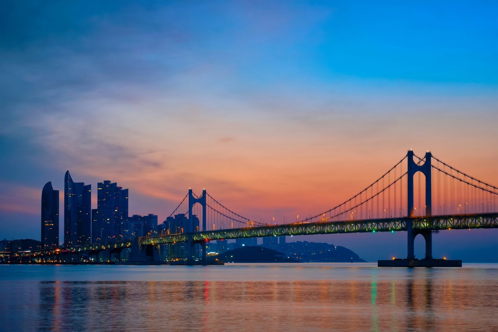 A suspension bridge and a group of towers stand in silhouette over calm water under an orange and blue sky