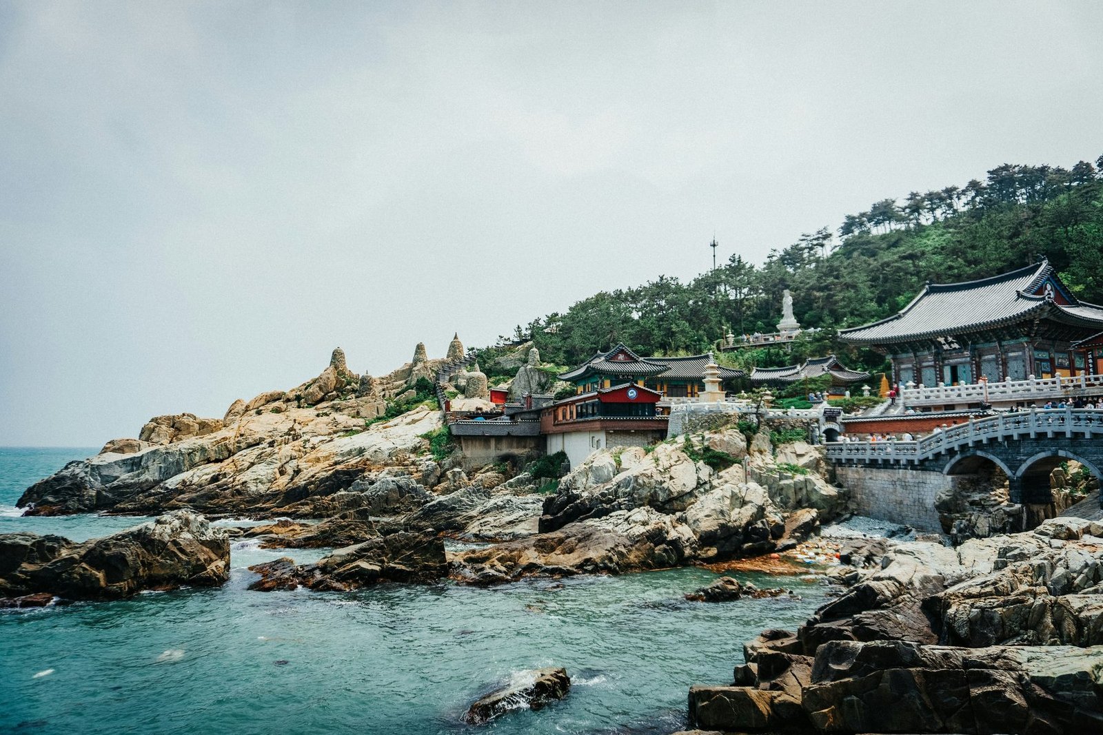 Temple halls with tiled roofs and a stone bridge sit on pale rocks above green sea water, with wooded hills behind