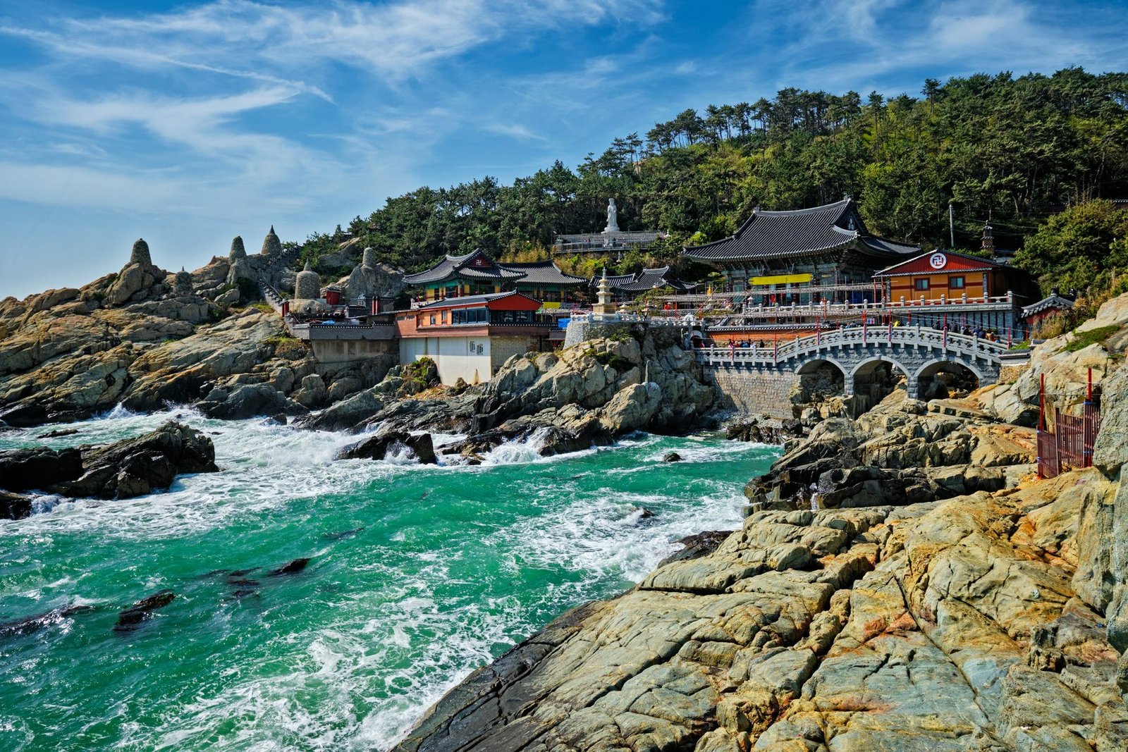 Temple halls with curved tiled roofs and a stone arch bridge sit on rocks above green surf below a wooded hill