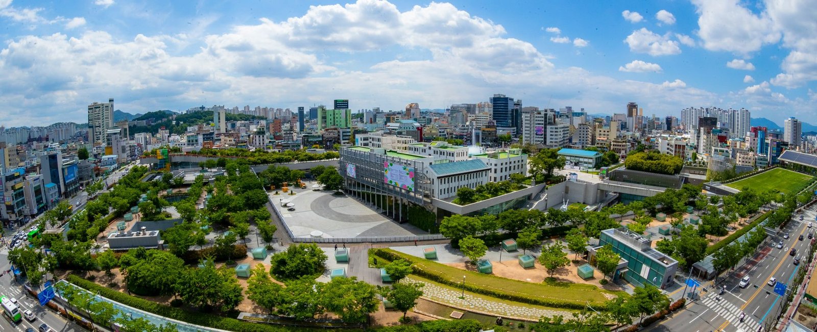A panoramic view of a large low complex with green roofs and sunken courts set among trees, ringed by city buildings and hills under clouds