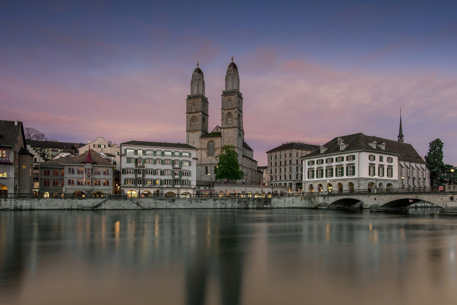 A twin-towered church and riverside buildings stand above smooth water under a violet sky