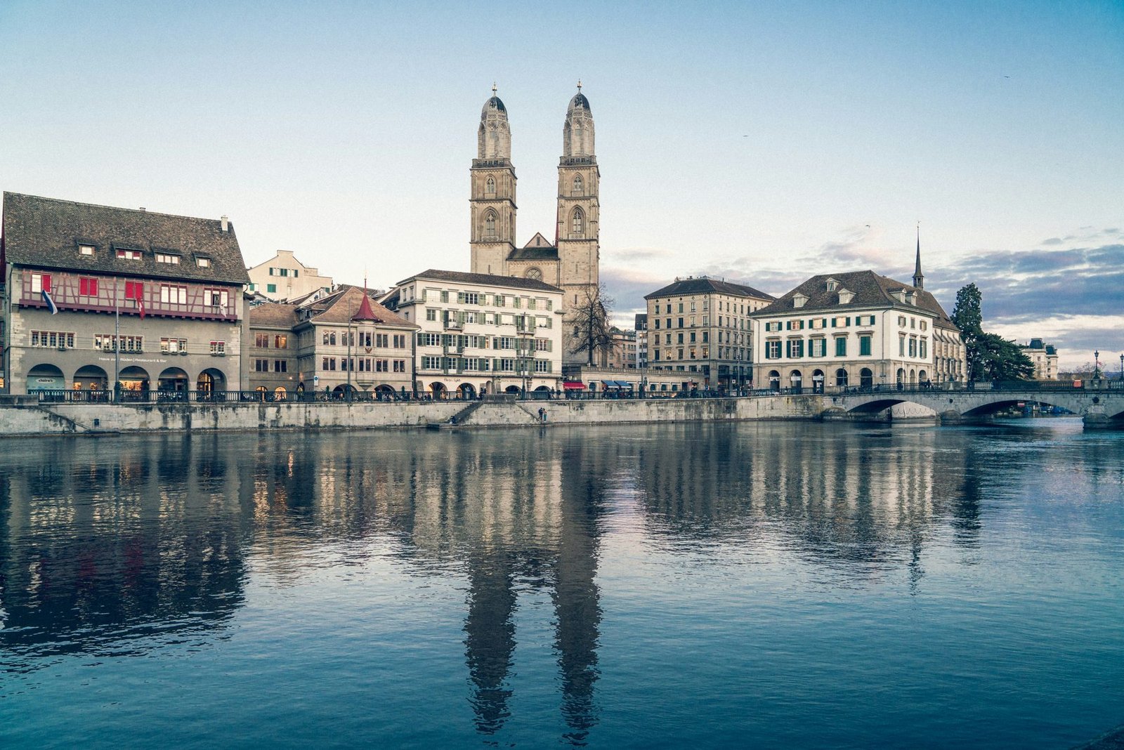A church with two domed towers rising behind riverside guild houses, all reflected in calm water