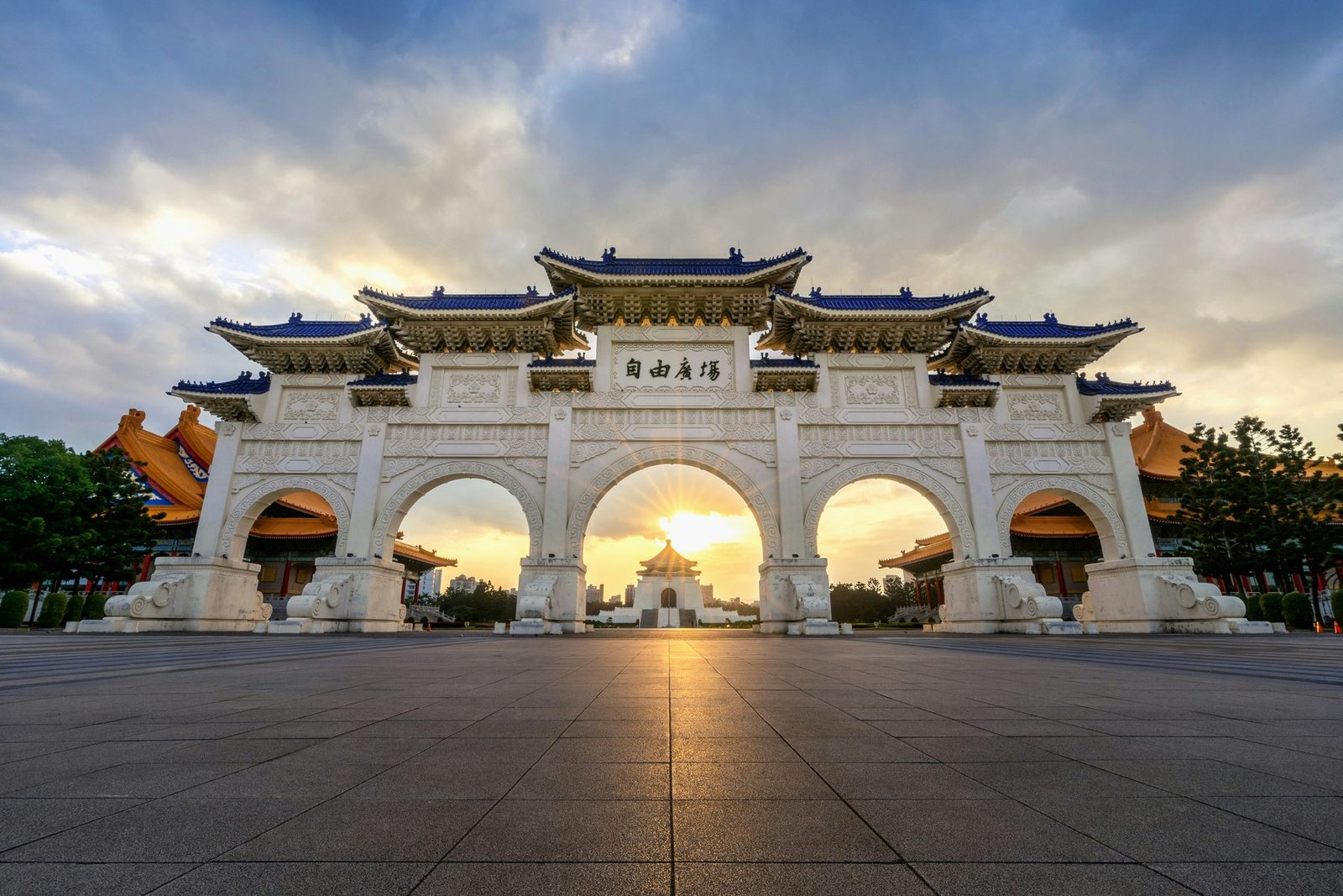A white five-arched gate with blue-tiled roofs stands at the edge of a wide paved square, with the sun setting through its central arch
