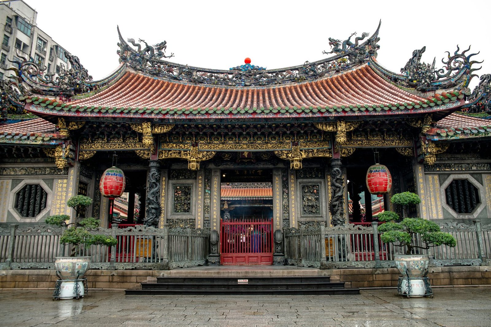 A temple hall with a sweeping tiled roof crowned by dragon figures stands behind a red gate, red lanterns and potted pines on wet paving