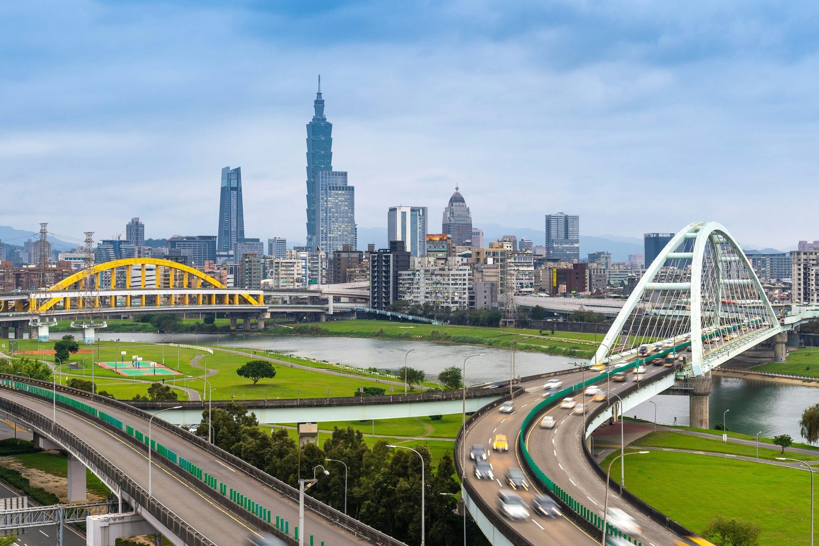 Curving elevated roads and two arched bridges, one yellow and one white, cross a river park with a tall tower and the city skyline behind