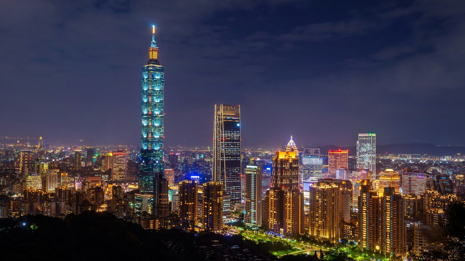 A tall tiered tower lit in blue-green stands above lit high-rises under a dark purple sky
