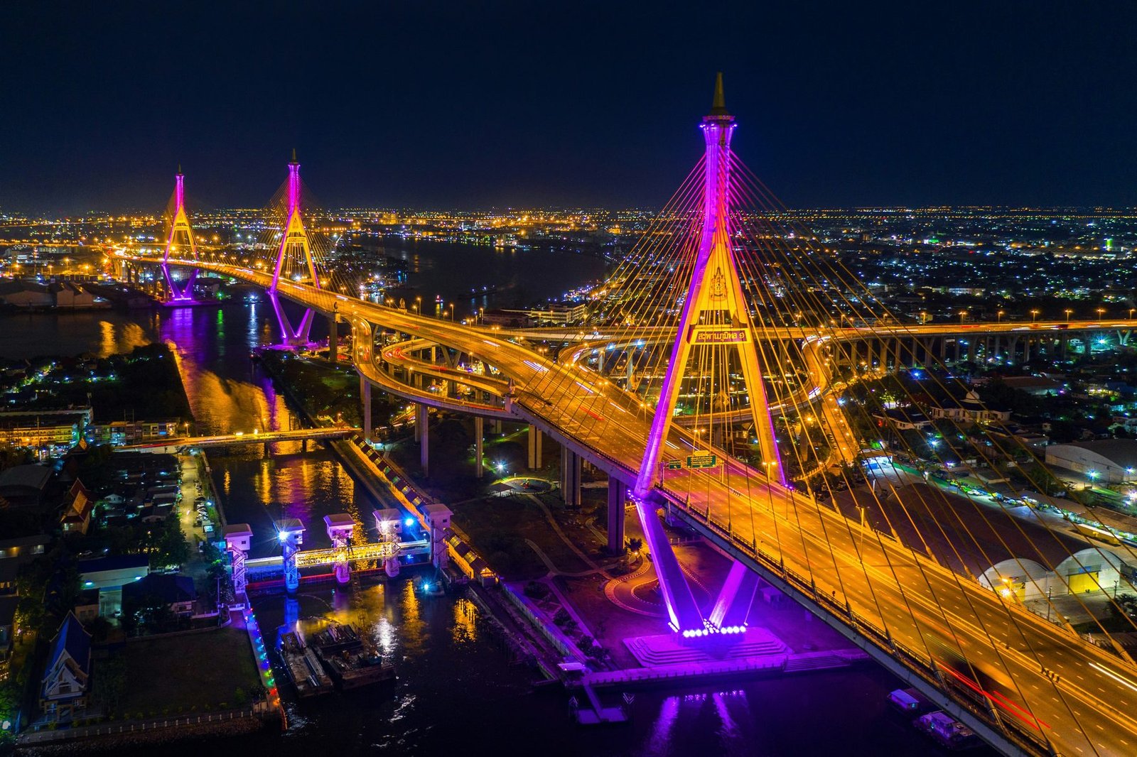 Two cable-stayed bridges with diamond-shaped masts lit in magenta and gold curve over a river and a looping interchange