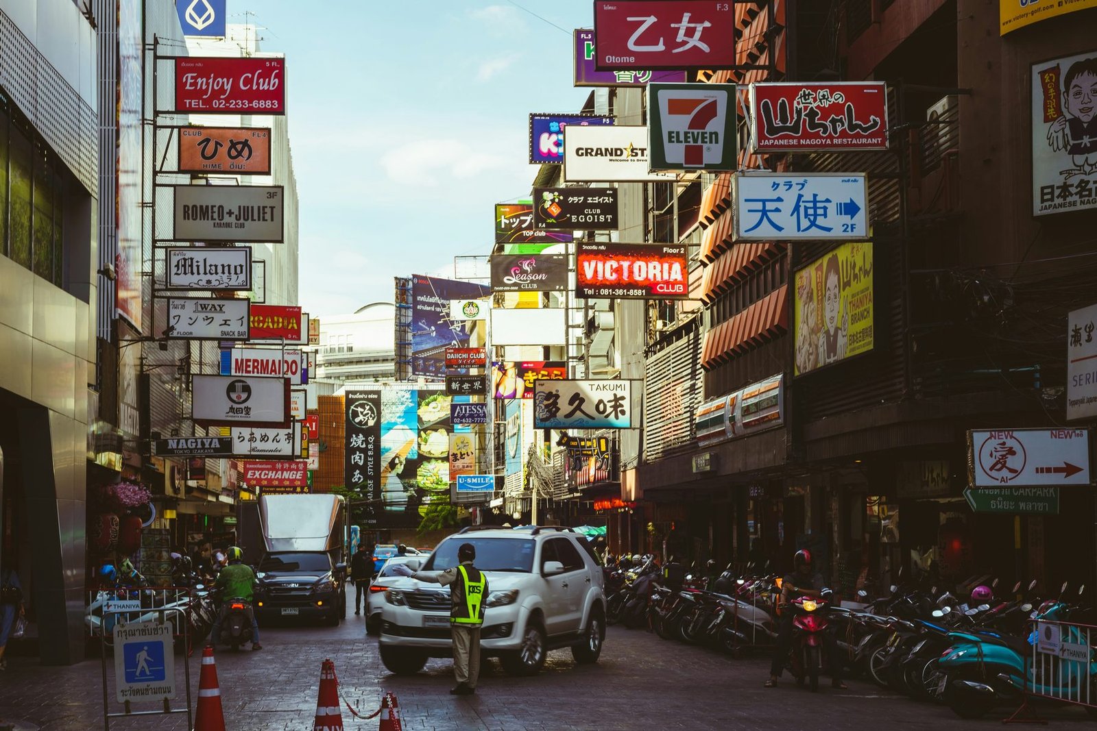 A narrow street crowded with overhanging shop and bar signs in several languages, with a white car, parked motorbikes and a traffic officer