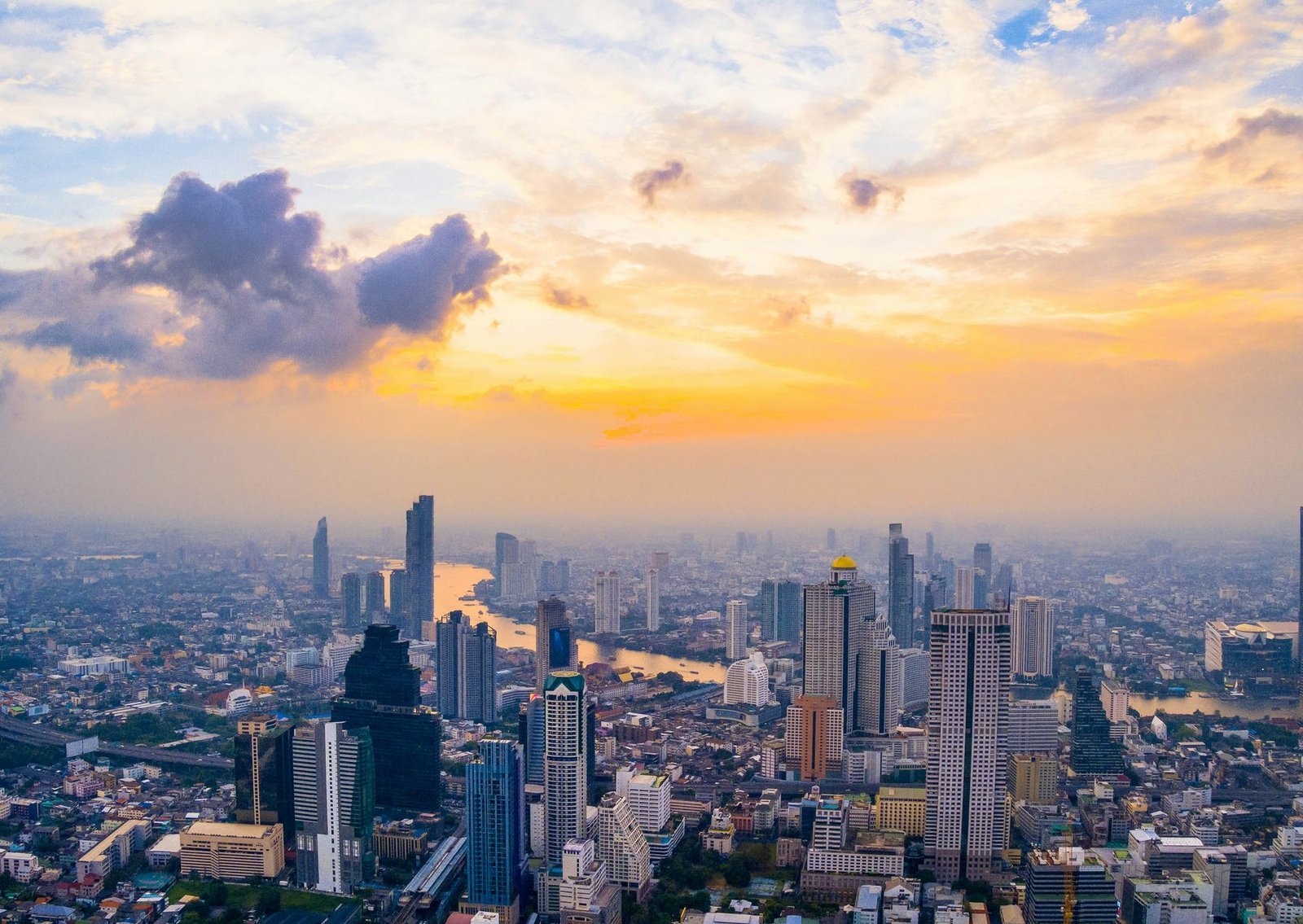 High-rise towers stand in blue haze around a bend of river under a yellow and orange sky with dark clouds