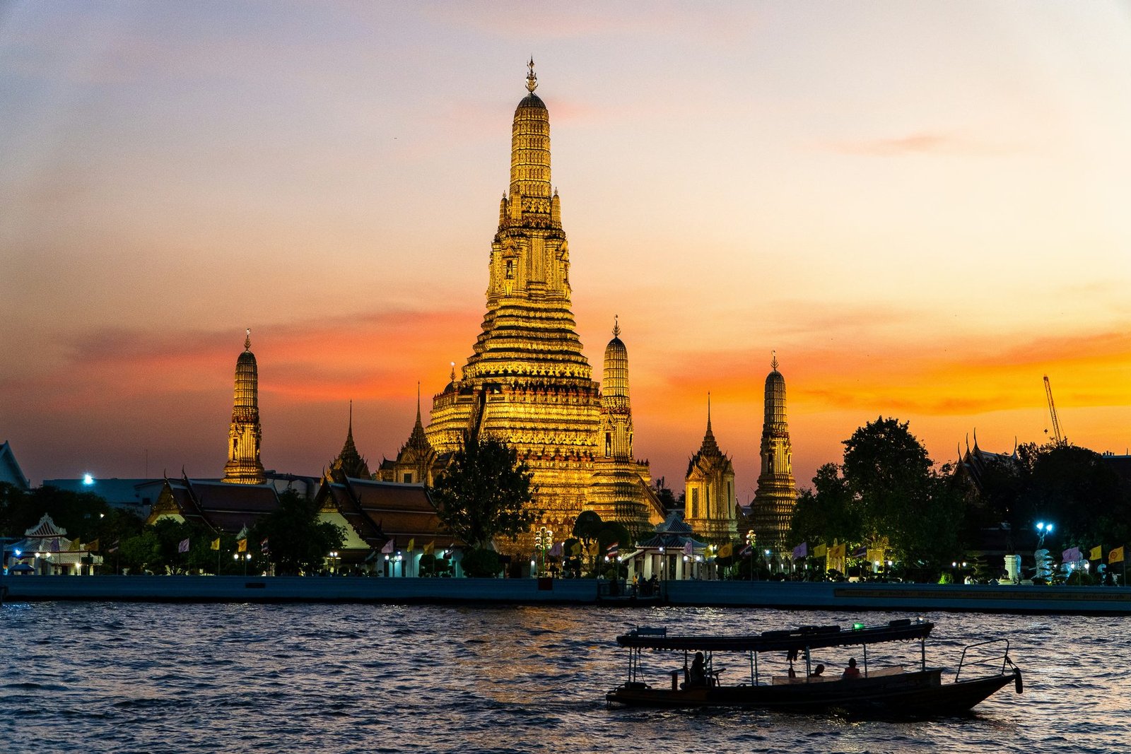 A floodlit temple spire and its smaller towers stand against an orange sky as a long narrow boat crosses the water in front