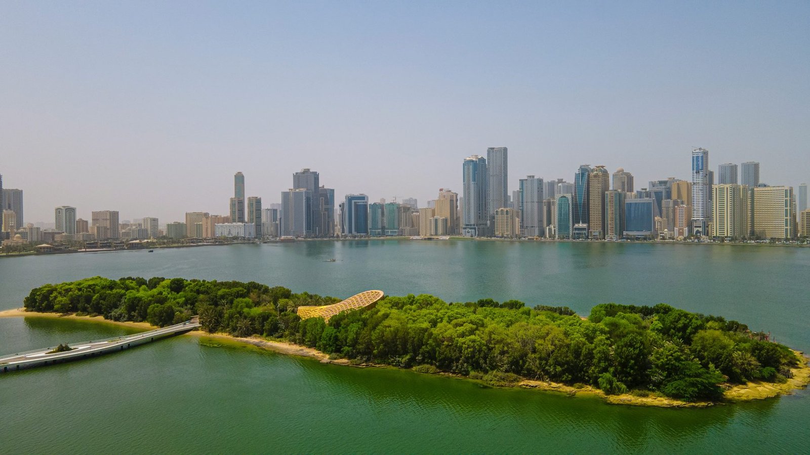 A small green island with a footbridge sits in a calm lagoon, with a line of towers on the far shore