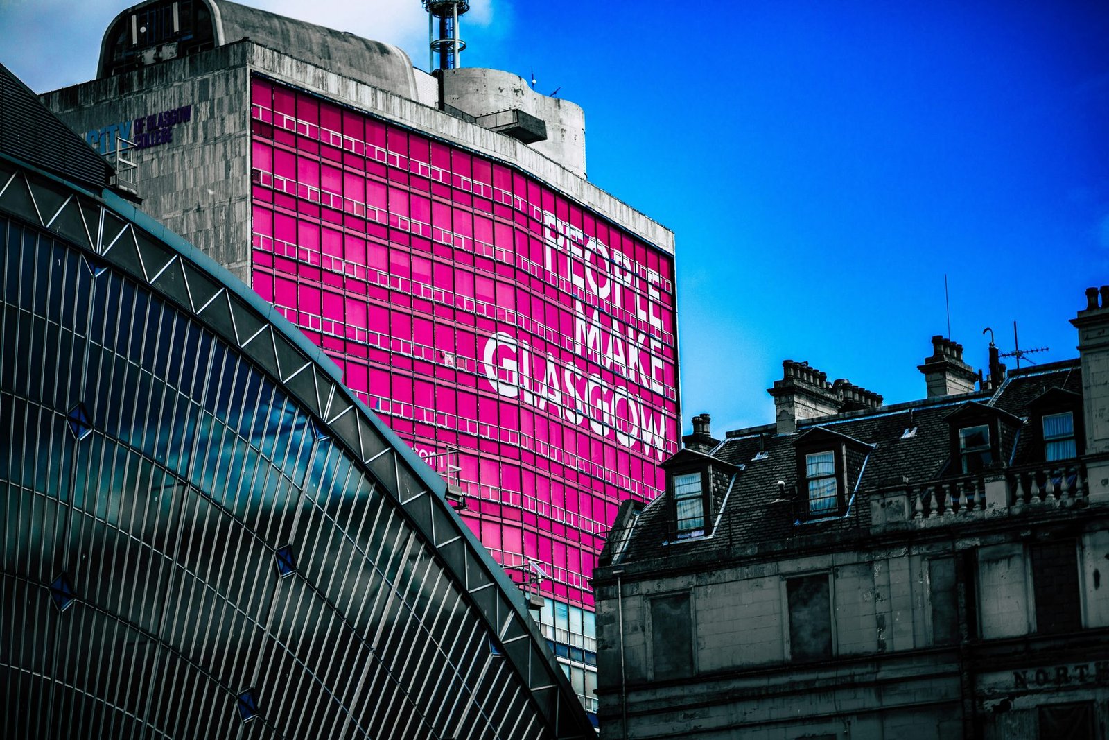 A tower block wrapped in bright pink with the words 'People Make Glasgow', seen over a curved glass station roof against a deep blue sky