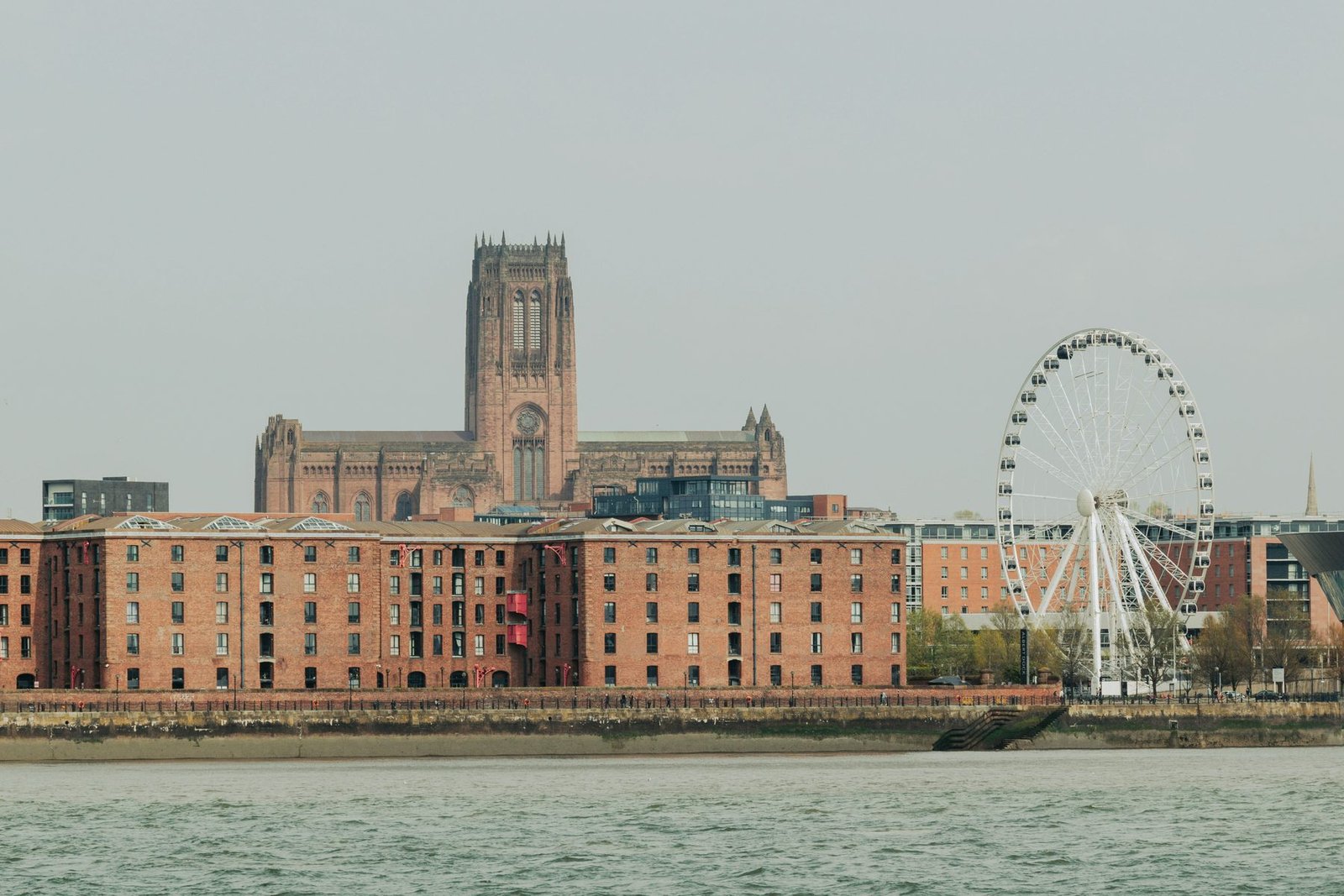 A large sandstone cathedral tower rising behind long red-brick dock warehouses and an observation wheel, with gray river water in front