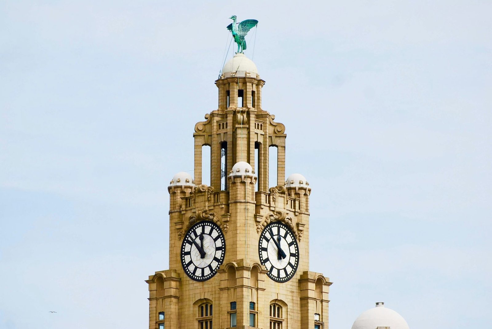 A stone clock tower with two clock faces topped by a green copper bird statue against a pale sky