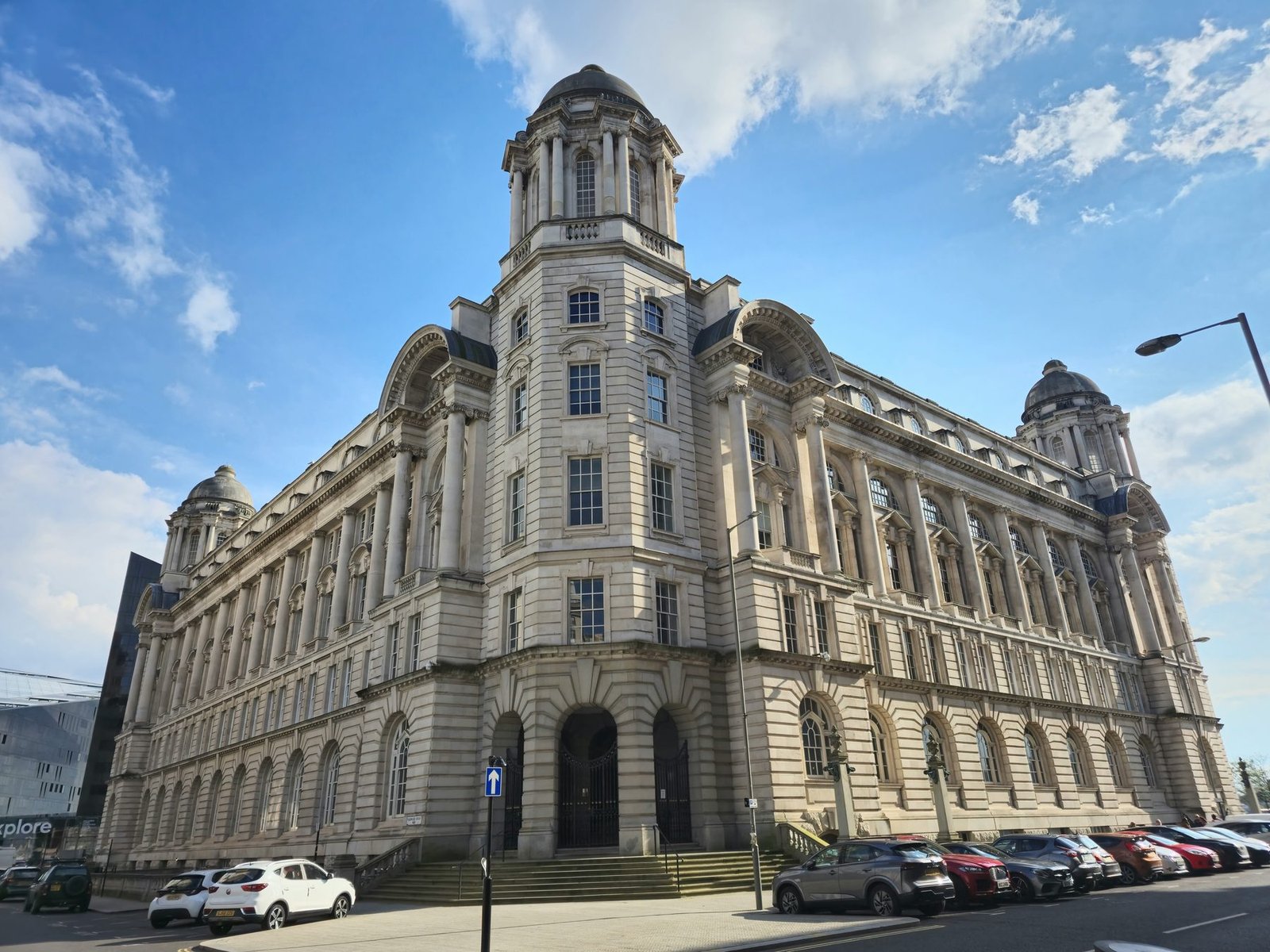 A large pale stone Edwardian building with a central dome and corner towers, seen from street level with parked cars in front