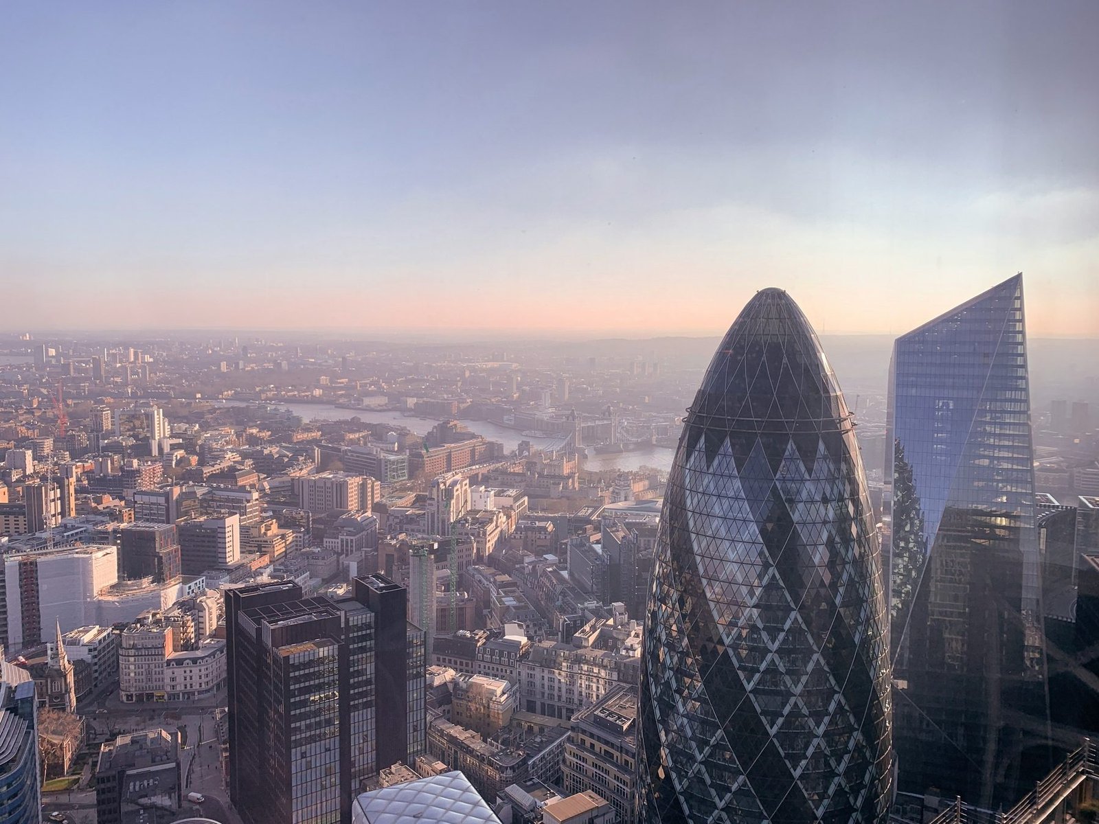 A rounded glass tower with a diamond lattice rises beside an angular skyscraper above a hazy city under a pink and blue sky