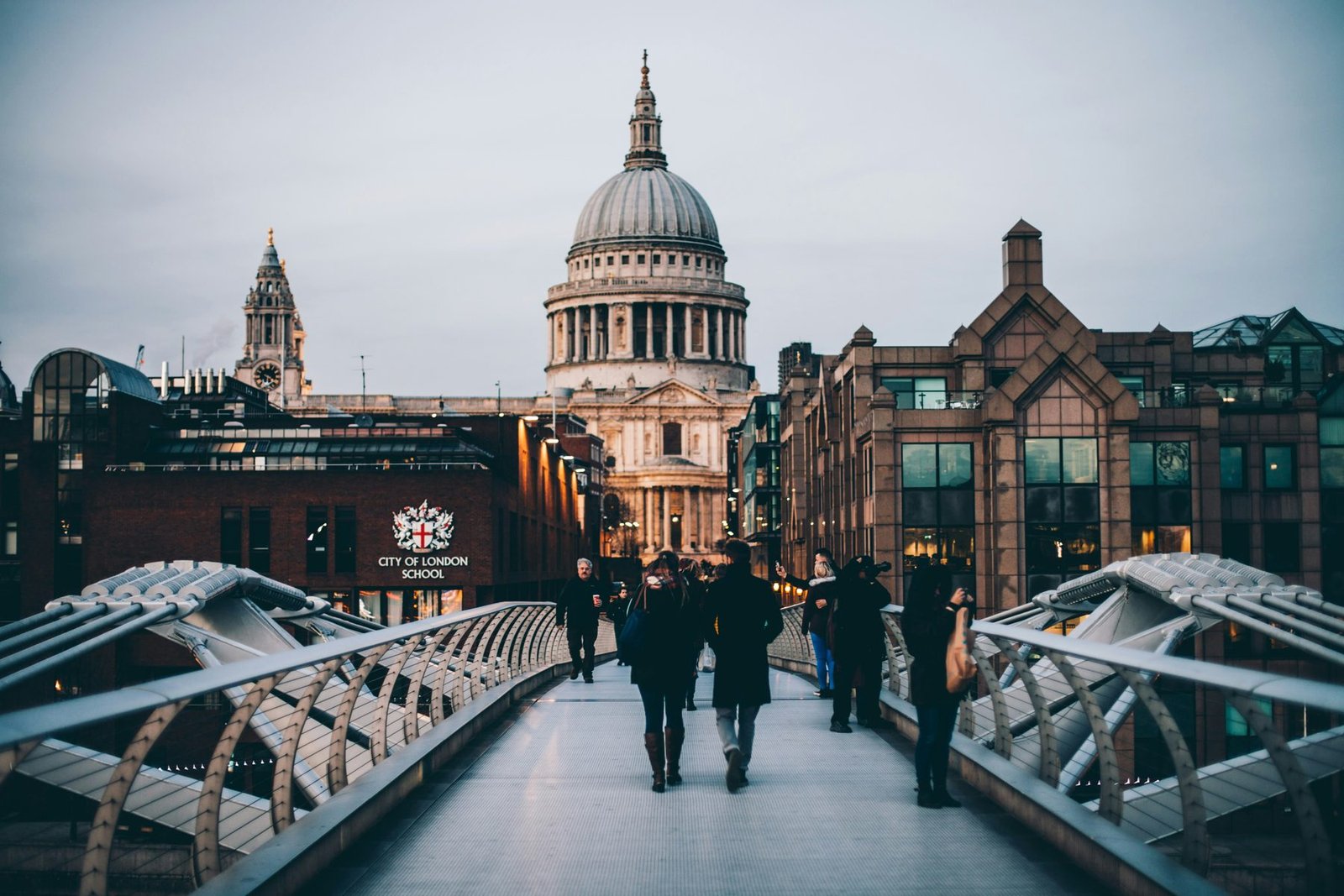 Pedestrians walking along a steel footbridge toward a large domed cathedral framed by brick and glass buildings