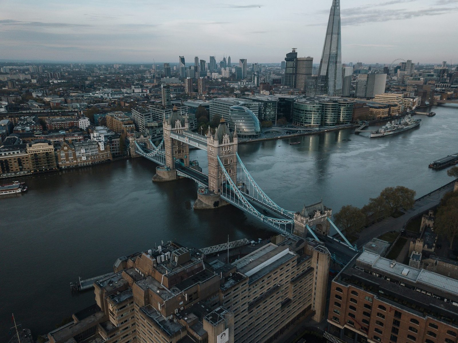 Aerial view of a twin-towered bascule bridge across a wide river, with a tall glass spire and office blocks on the far bank at dusk