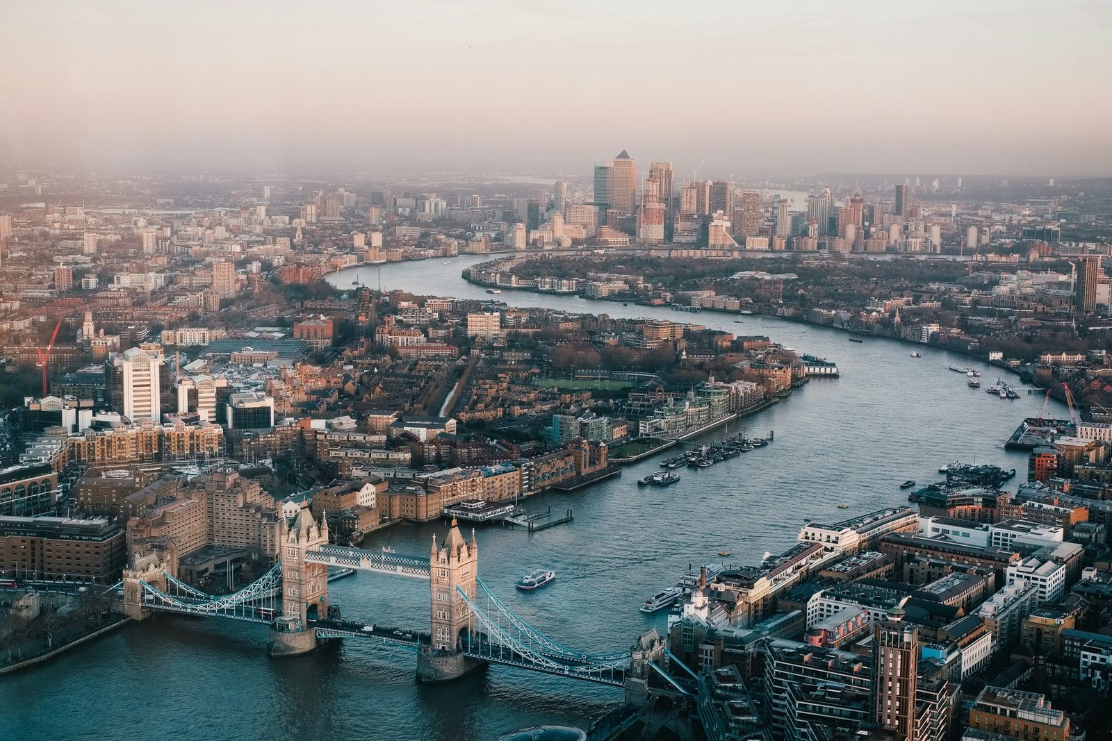 Aerial view of a twin-towered bridge and a river curving east through the city to a distant cluster of towers in warm low light