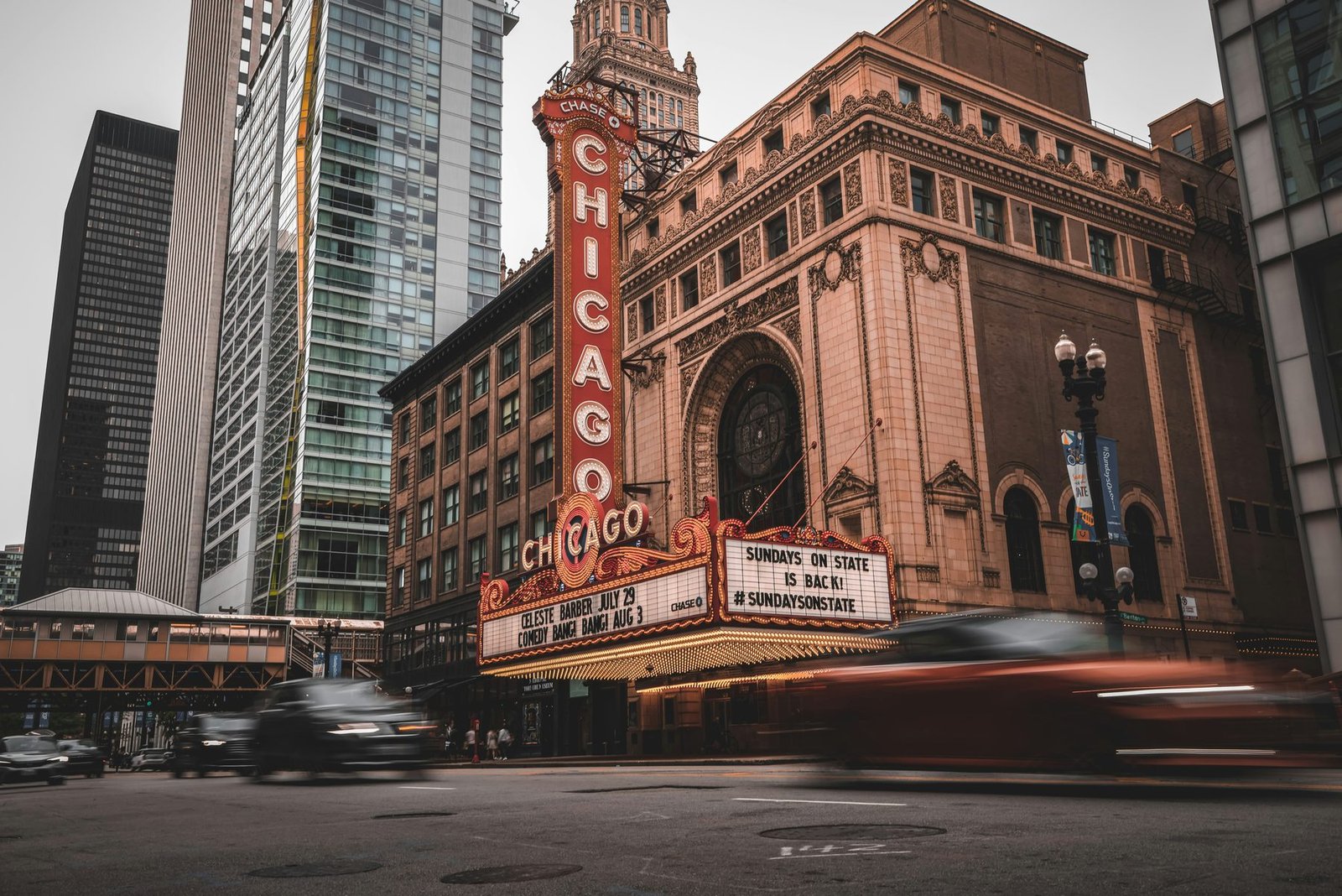 A vertical red sign spelling CHICAGO rises above a theater marquee on an ornate terracotta facade, with cars blurred in the street