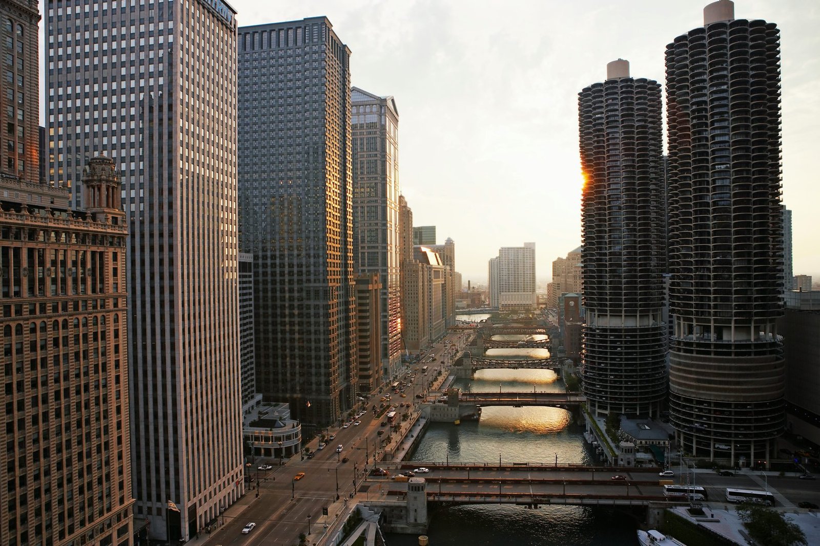 Two round towers with scalloped balconies stand beside a river crossed by a line of bridges, with the sun low between the buildings