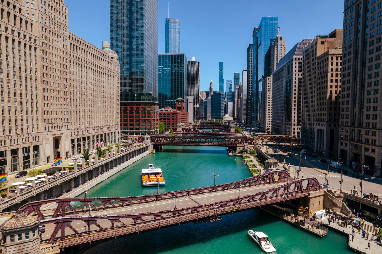 A green river with tour boats passes under a red steel bridge between a large stone building and glass towers