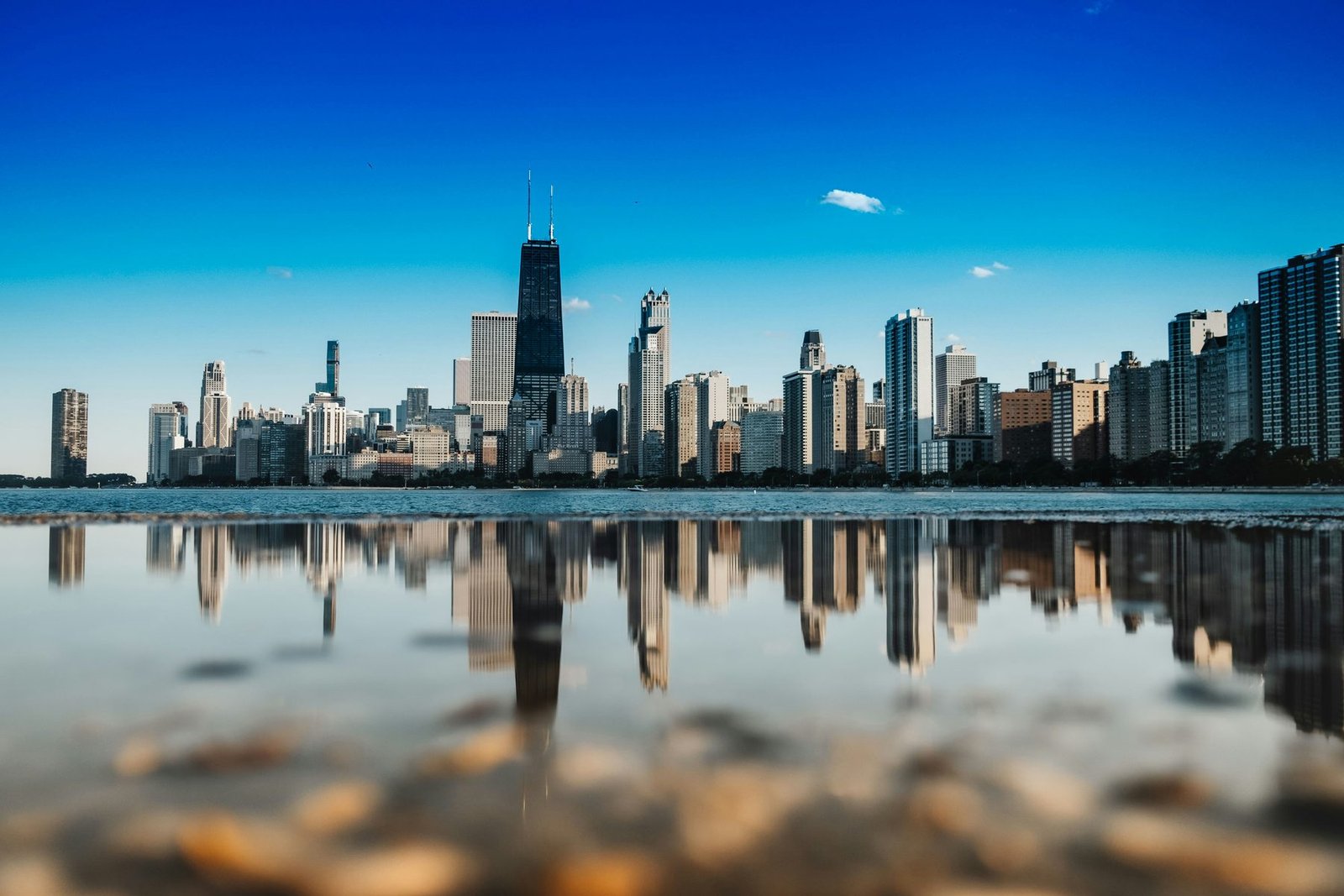A row of towers along a lakeshore is mirrored in still water in the foreground under a deep blue sky
