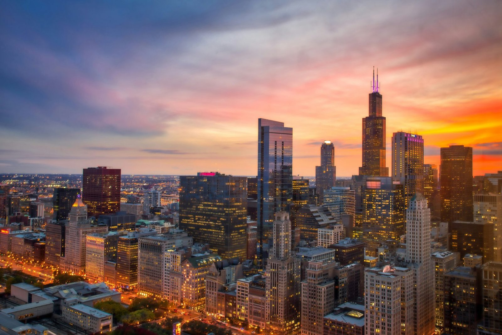 Dense high-rises with lit streets stand beneath a tall dark tower with twin antennas under an orange and purple sky