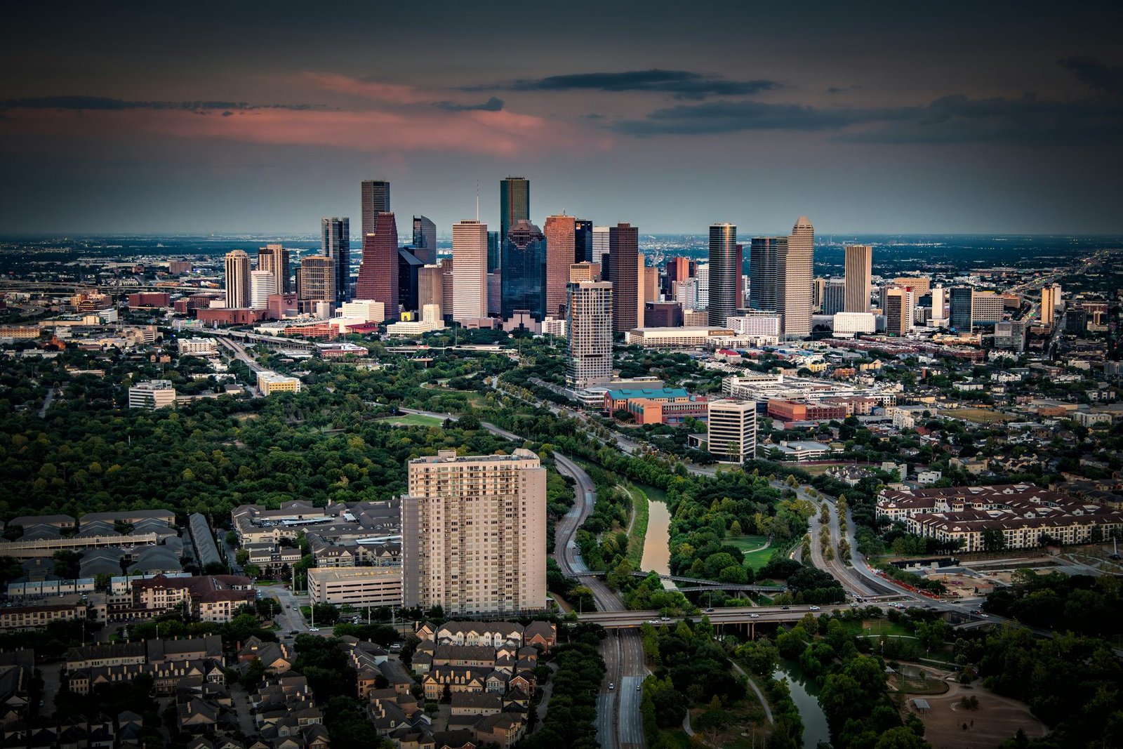 A cluster of towers sits beyond a wooded parkway and winding roads under a dark sky with a pink band of cloud