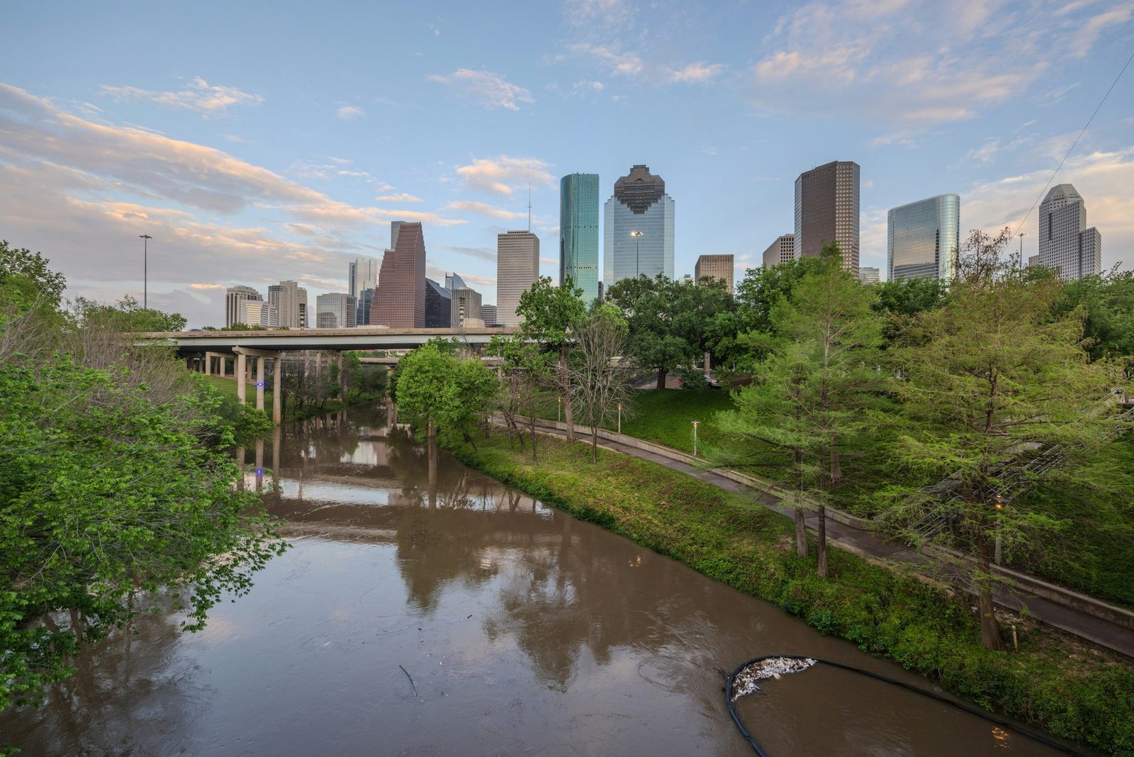 A brown stream winds between green banks and under road bridges toward a group of glass towers