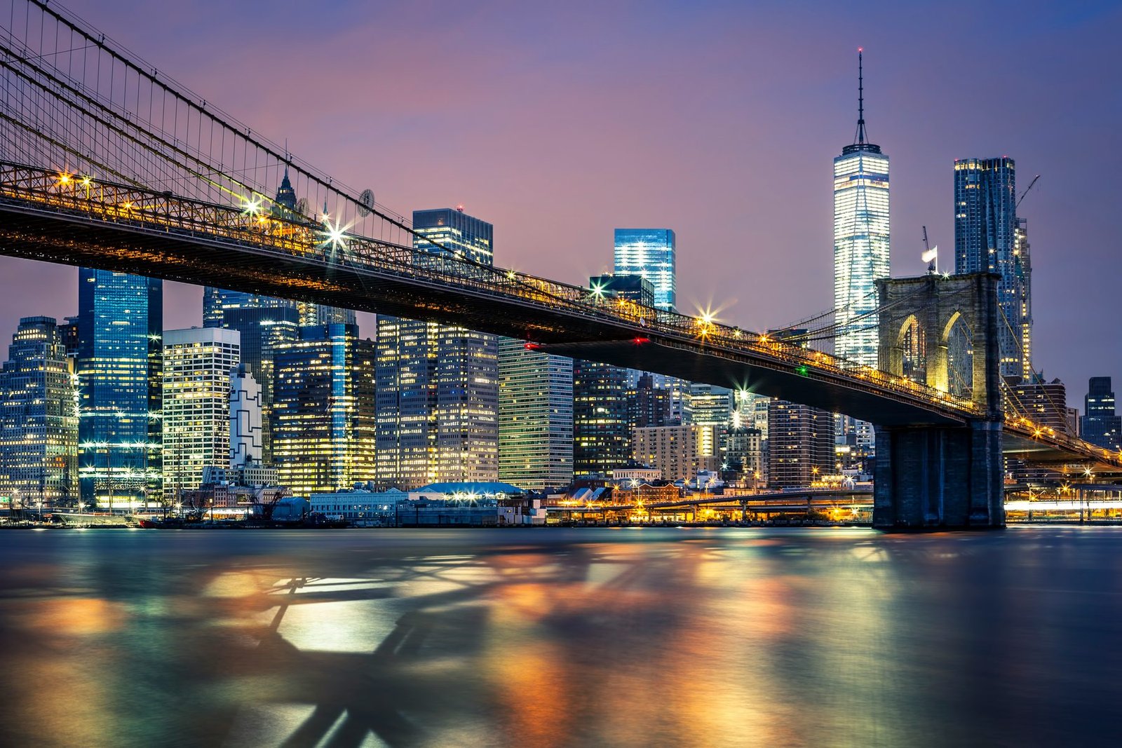 A lit suspension bridge crosses a river toward illuminated skyscrapers under a purple night sky, with light smeared on the water
