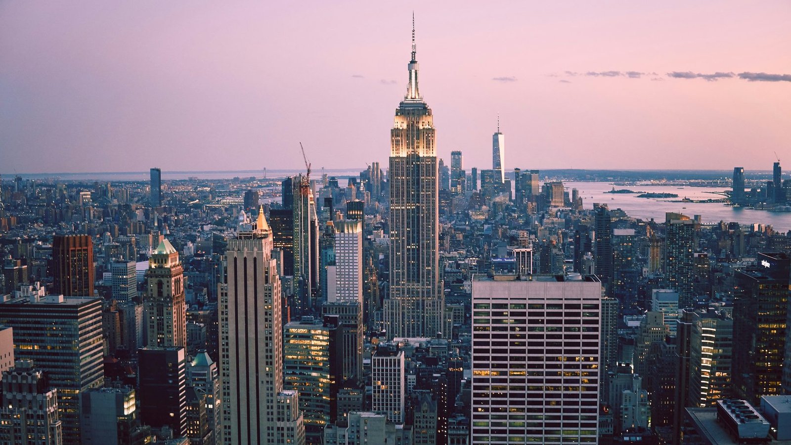 A tall spired skyscraper stands above a wide cityscape under a pink evening sky, with distant towers and water beyond