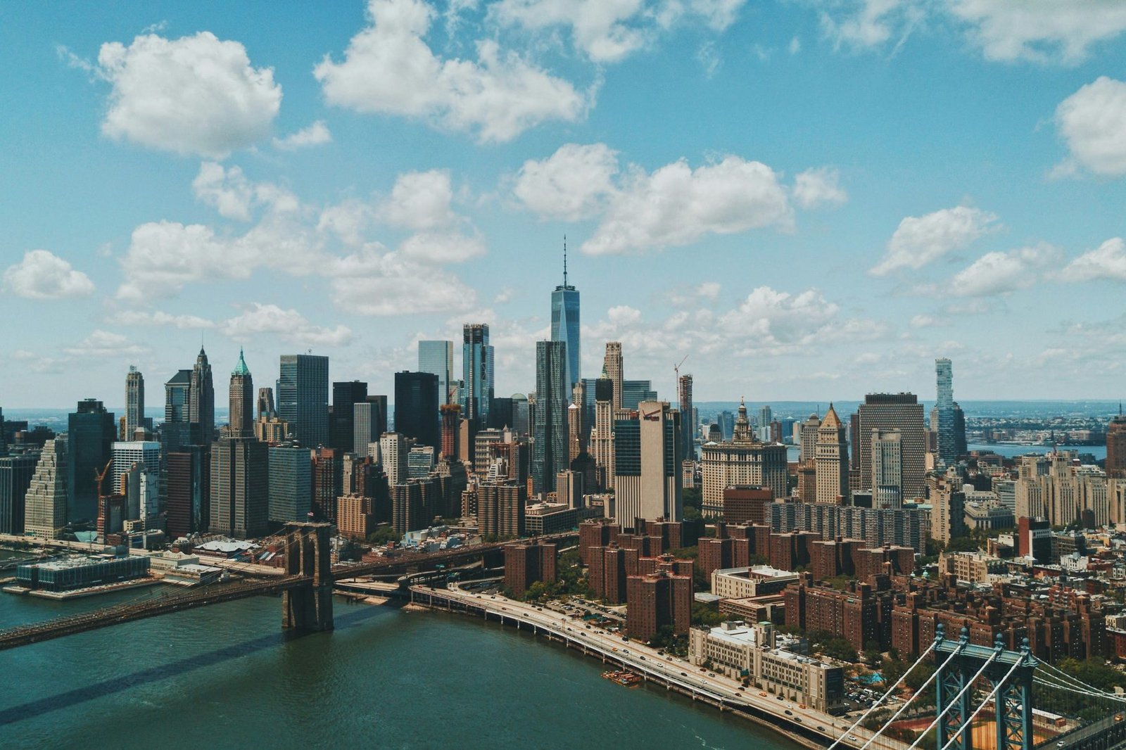 An aerial view of a dense cluster of skyscrapers at the tip of an island, with two suspension bridges crossing the river in the foreground